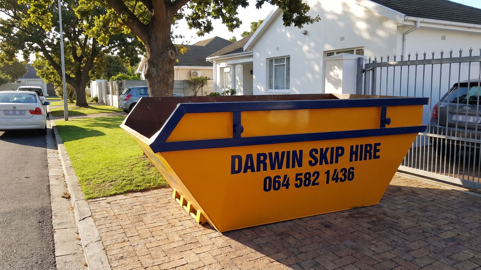 Empty amber Darwin Skip Hire mini skip on a paved driveway under oak trees.