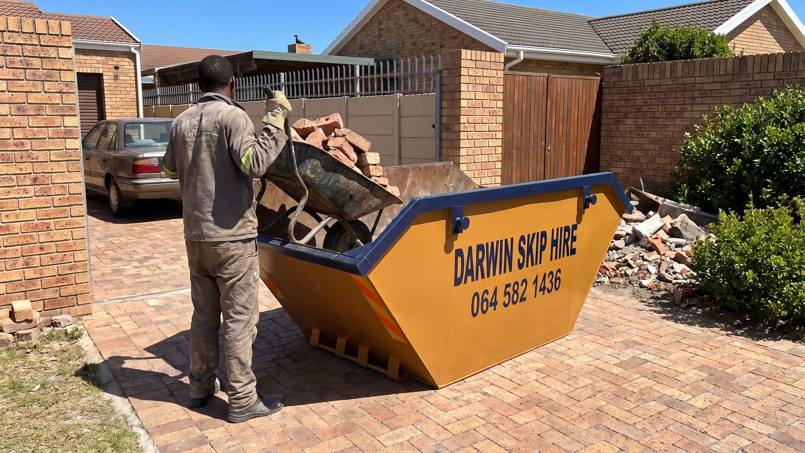 Worker tipping a wheelbarrow of broken bricks into an amber Darwin mini skip.