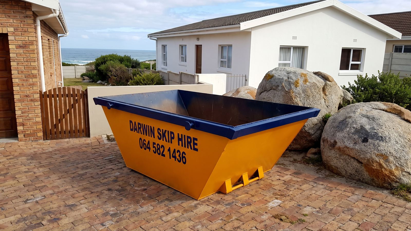 Amber Darwin mini skip on paving beside a white bungalow, granite boulders and the sea.