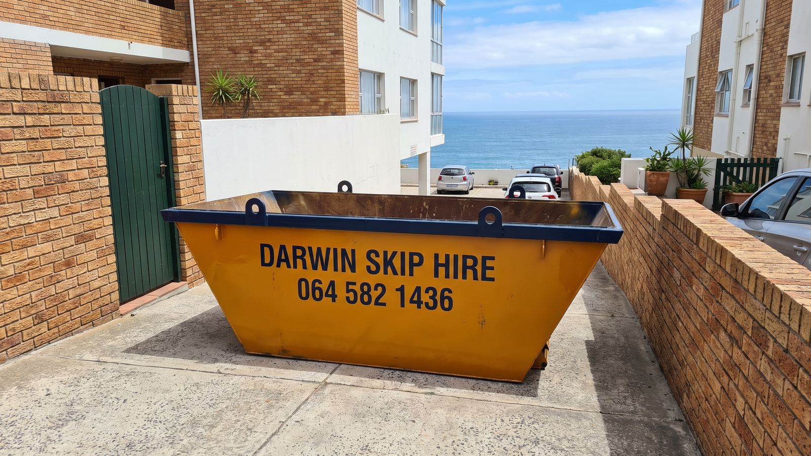 Amber Darwin mini skip on a narrow paved parking area between buildings above the sea.
