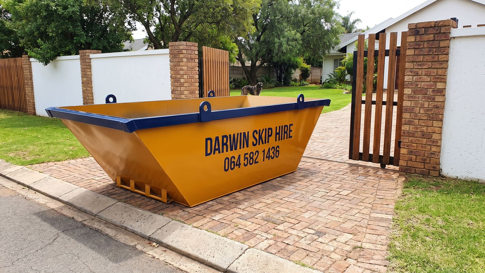 Amber Darwin mini skip on a brick driveway inside the open wooden gate of a large walled property.