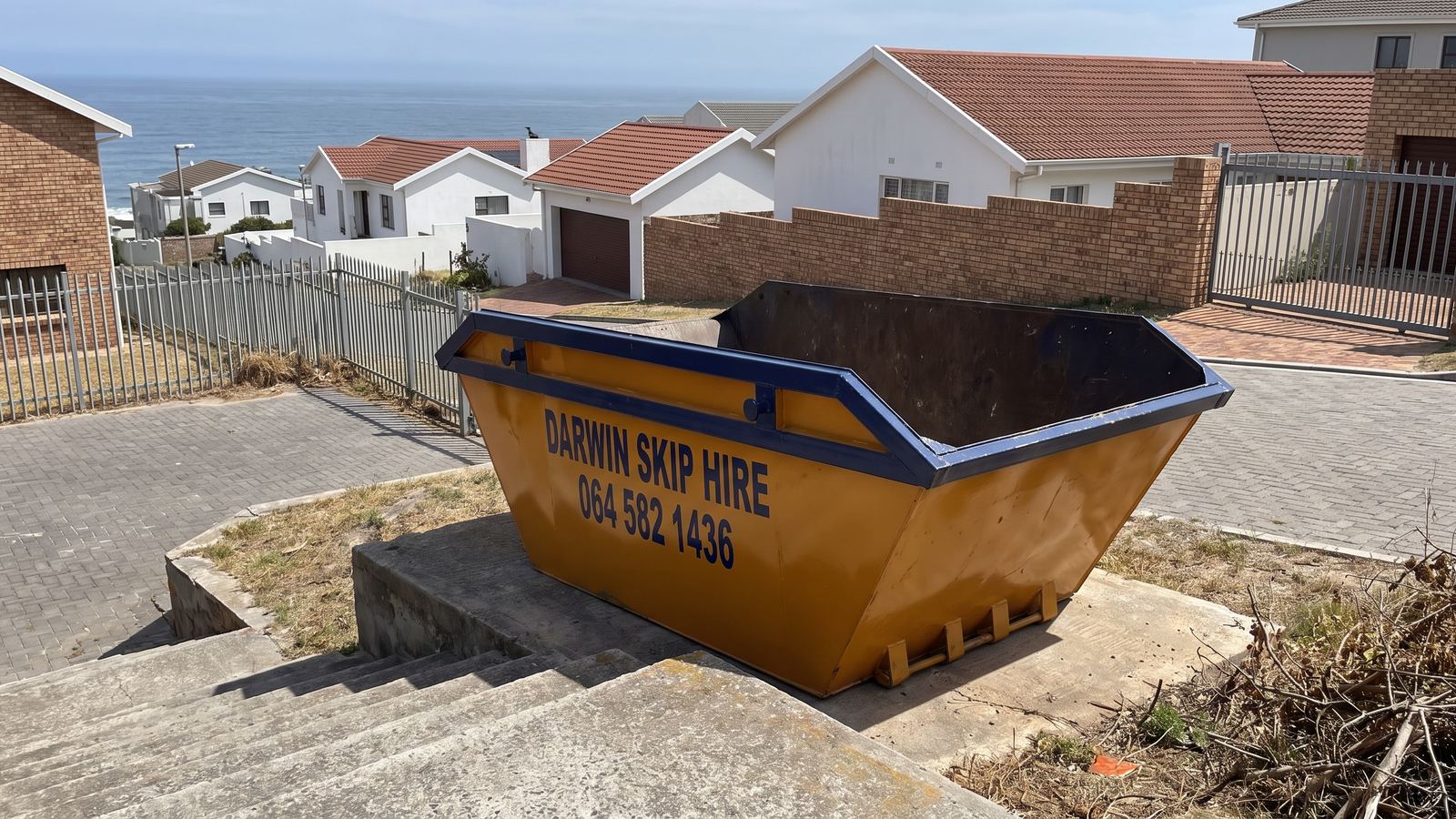 Amber Darwin mini skip on a concrete landing at the top of steps, with houses and the sea below.