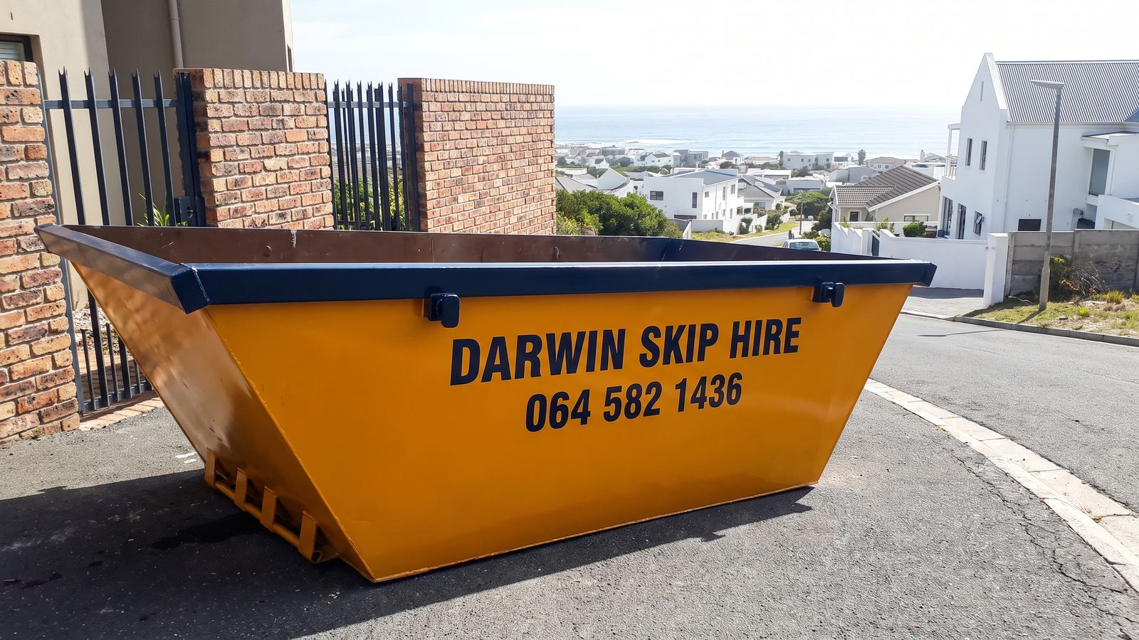 Amber Darwin mini skip on a sloping road beside a brick wall with the sea beyond.