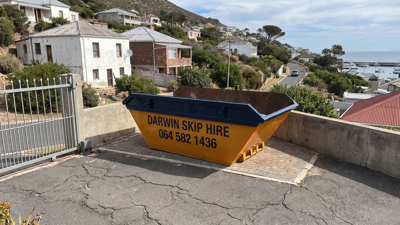 Amber Darwin mini skip on a small paved terrace above a mountainside road and a harbour.