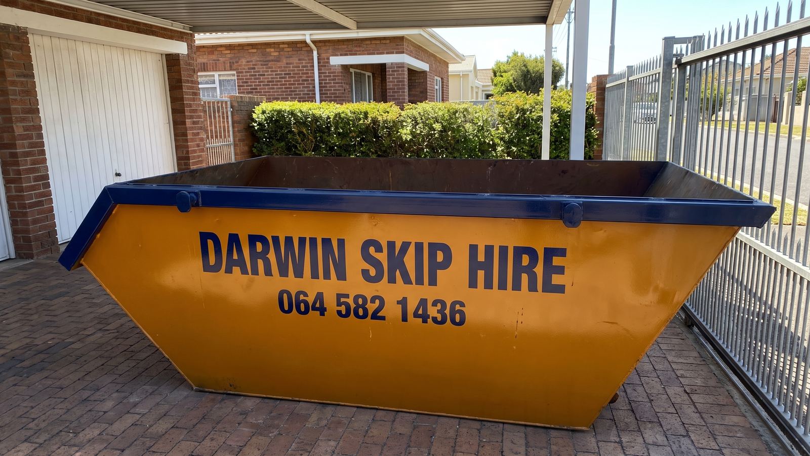 Amber Darwin mini skip on a brick driveway under a carport beside a face-brick house.