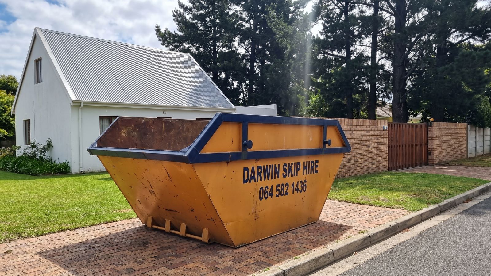Amber Darwin mini skip on a brick driveway beside a lawn and a cottage-style house with tall pines.