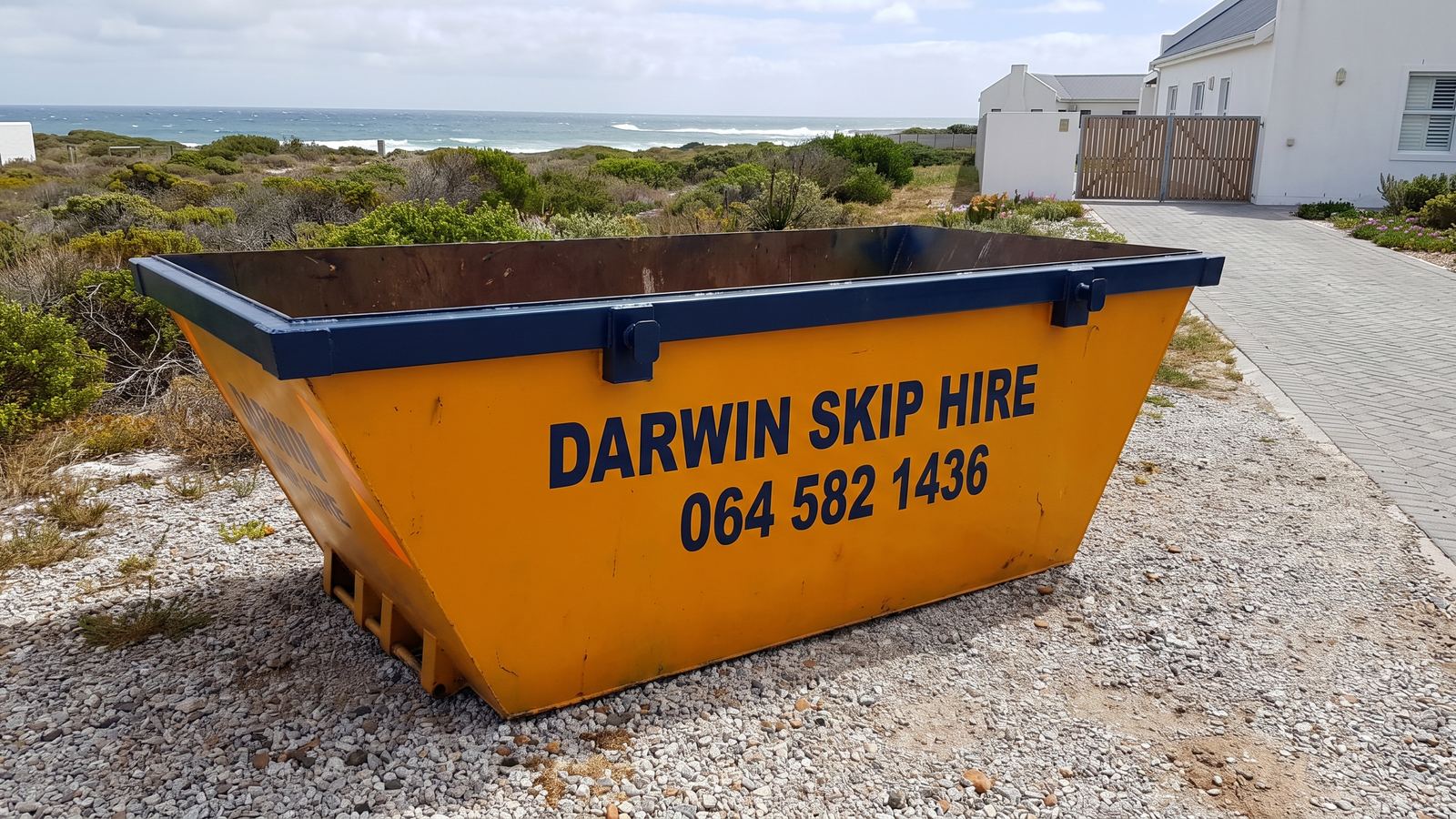 Amber Darwin mini skip on a gravel plot in coastal fynbos with the sea beyond.